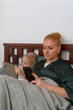 Mother and daughter are relaxing in bed under a blanket, looking at their individual smartphones and tablets, using technology for communication and entertainment