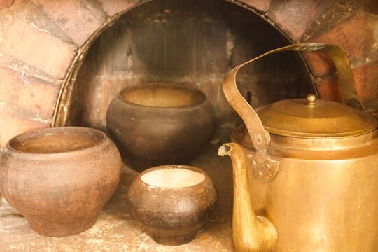 Vintage metal kettle surrounded by rustic clay pots inside an old brick oven or fireplace. Warm golden light highlights the aged surfaces and traditional cookware, creating an atmospheric rural stil