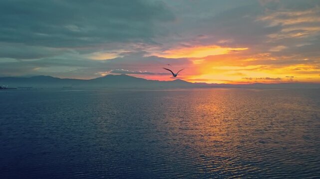 KALOCHORI, GREECE: Cinematic aerial video panning right across a tranquil lagoon at sunset, featuring vibrant colorful clouds reflecting on the calm water, flying birds, and mountain silhouettes in th