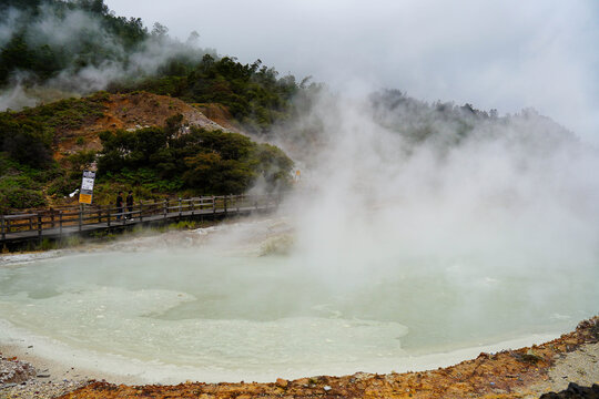 Steaming geothermal hot spring lake with wooden walkway and lush volcanic hillside