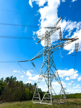 High voltage power line tower against blue sky. A tall steel transmission pylon stands in a field near green trees under bright daylight.
