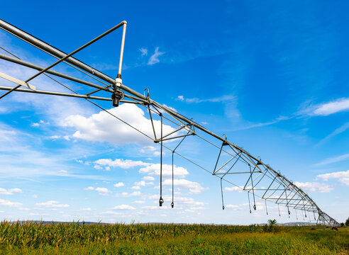 Long pivot irrigation pipe spanning green agricultural land. Steel truss line extends across farmland towards horizon under sunlight and blue sky with clouds.