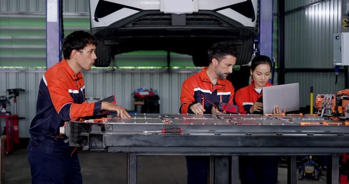 EV diagnostic teamwork in modern workshop: three technicians operate handheld tester and laptop beside exposed battery pack, coordinating inspection during electric vehicle maintenance service.