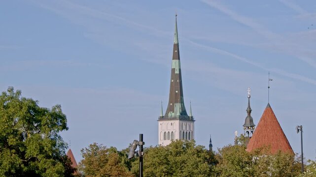 Locked 4K footage of St. Olaf&rsquo;s Church spire rising above the medieval towers and green trees of Tallinn Old Town, Estonia. Historic city skyline under a clear blue summer sky.