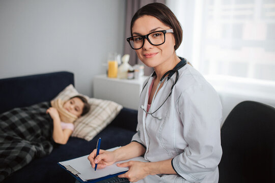Nice young female doctor look straight on camera and smile. She sit besides sick small girl. Child lying on bed and covered with blanket.