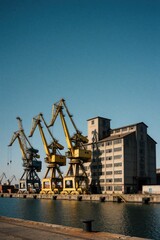 Fototapeta premium Industrial harbor with yellow gantry cranes, large concrete warehouse by water. Heavy machinery stands at shipyard pier under clear blue sky. Maritime logistics terminal for international trade,