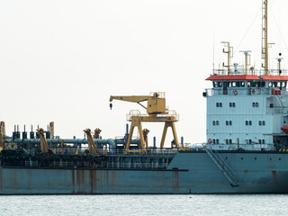 Large dredger ship with yellow deck crane, pipes and white bridge with orange roof anchored on calm river, rusty steel hull and industrial marine equipment under hazy sky © Tavan