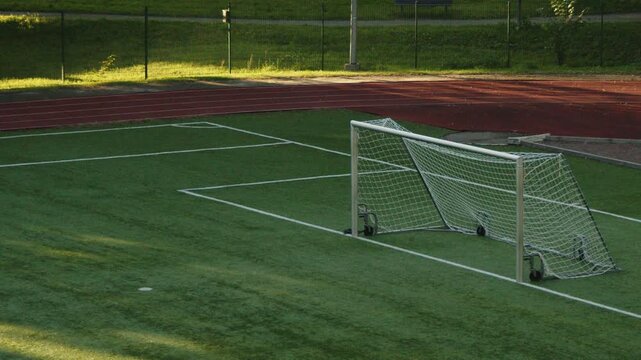 Locked 4K footage of an empty football field with a portable soccer goal, artificial turf and running track in Tallinn, Estonia. Quiet outdoor sports facility illuminated by warm evening sunlight.