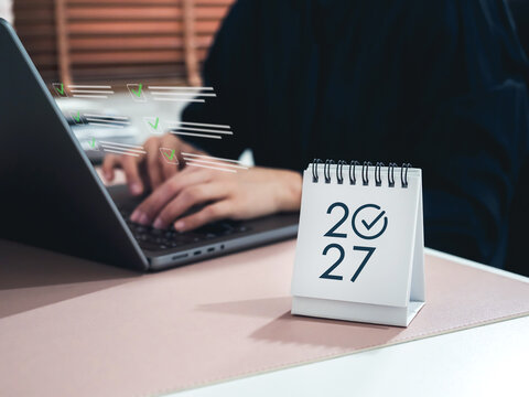 Desk calendar marked 2027 beside businessperson typing on laptop with digital checklist icons. Annual business planning, goal achievement, task management, and strategic execution for the year ahead.