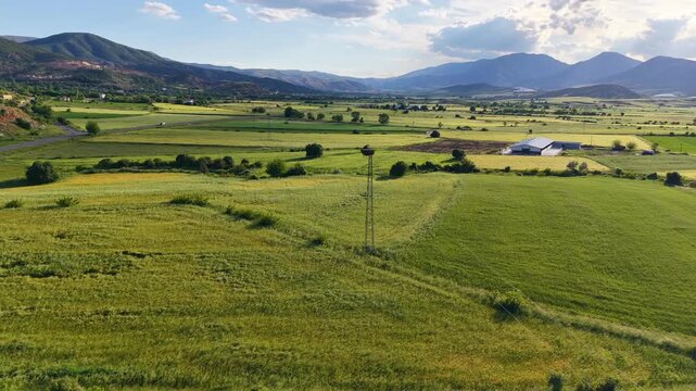 Smooth slow motion aerial orbit around a stork nest and power pole, revealing a lush green valley, mountains and a scenic lake as the view gradually changes.