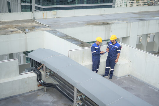 Rooftop engineer technician inspection maintenance hardhat safety ventilation team checking duct and pipe system on commercial building roof, using tablet for facility management, cautious focus