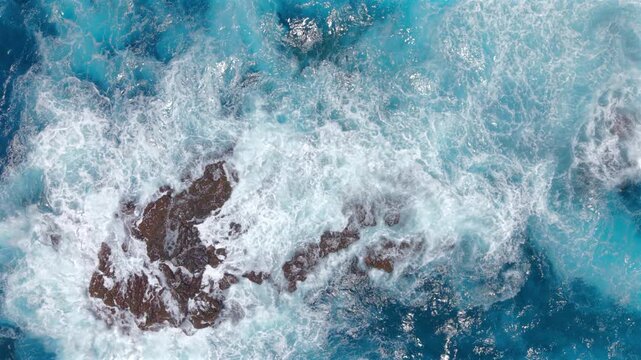 Aerial top down view of turquoise ocean waves breaking into white foam over dark volcanic rocks at Kaihalulu Beach on Maui Hawaii island with swirling currents and vivid blue water.