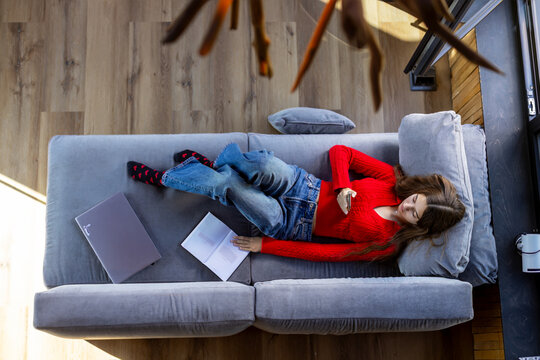 Woman Relaxing on Sofa with Smartphone, Book and Laptop. Overhead view of a woman in a red sweater relaxing on a gray sofa with a smartphone, open book, and laptop beside her.