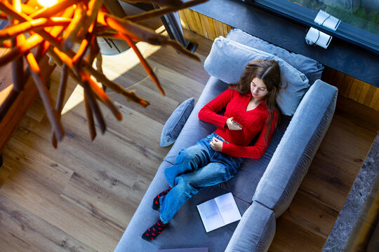 Young Woman Relaxing on Sofa and Using Smartphone. Young woman in a red sweater reclines on a gray sofa using her smartphone, with an open book beside her in a bright modern room