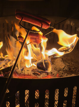 Delicious hot dogs and bacon roasting on metal skewers over a vibrant campfire in a backyard fire pit at night, capturing a cozy and rustic outdoor cooking experience.