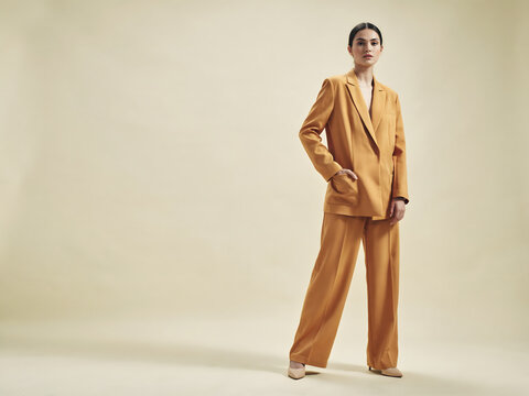 A woman posing in a mustard yellow oversized blazer and matching wide leg trousers with a hand in her pocket against a neutral studio backdrop