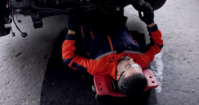 Adult male mechanic reaches upward with gloved hands while lying on creeper beneath electric vehicle, checking undercarriage parts during routine inspection inside modern automotive workshop.