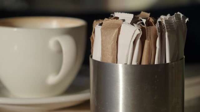 Close up view of sugar packets in metal holder near white coffee cup with latte art on table in cafe