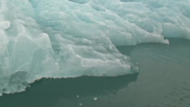 A large blue iceberg fills a close view above dark cold water. Additional visible detail includes surface texture and natural light, with natural light defining the surrounding forms.