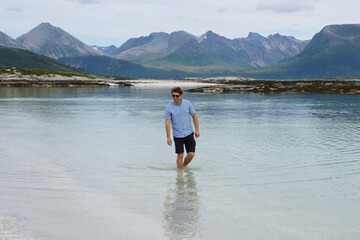 Young man walking barefoot in the crystal clear water of Sommaroy beach in Norway. Traveler enjoying summer vacation on the arctic coast of northern Norway. © vaitekune