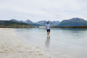 Young man walking barefoot in the crystal clear water of Sommaroy beach in Norway. Traveler enjoying summer vacation on the arctic coast of northern Norway. © vaitekune