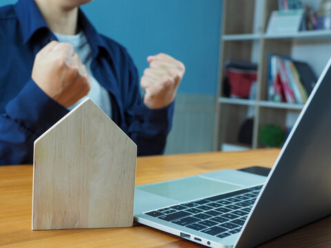 Wooden house model on desk beside laptop with person raising clenched fists in background expressing excitement and success, home office workspace scene