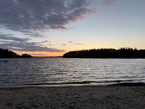 A serene Finnish lake at sunset with calm, rippling waters, mossy foreground rocks, and silhouetted pine forests under a dramatic golden hour sky.