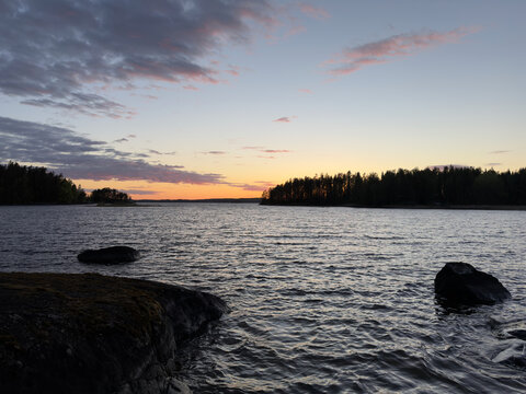 A serene Finnish lake at sunset with calm, rippling waters, mossy foreground rocks, and silhouetted pine forests under a dramatic golden hour sky.