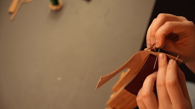 Female artisan hand stitches a leather accessory with green thread using needles. Traditional leathercraft process for handmade goods, fashion and small business