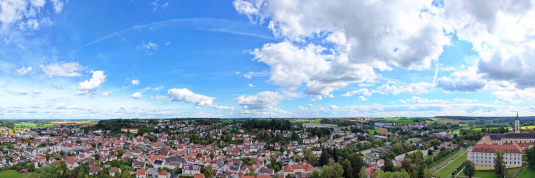 Weites Luftbild-Panorama von Ochsenhausen in Oberschwaben mit Blick &uuml;ber die Stadtstrukturen und Wohngebiete, umgeben von gr&uuml;ner Natur unter blauem Sommerhimmel.
