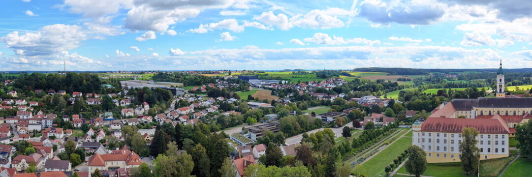 Malerische Luftaufnahme von Ochsenhausen mit der barocken Klosteranlage, eingebettet in die idyllische, gr&uuml;ne H&uuml;gellandschaft Oberschwabens unter blauem Sommerhimmel mit Wolken.