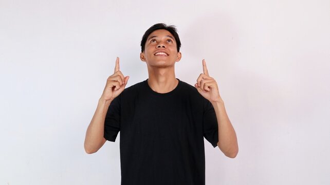 Young Asian man looking up with an amazed expression while pointing both index fingers toward the sky against a white background.