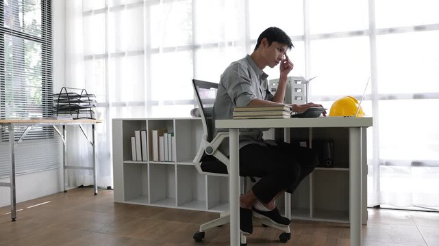 Overworked Asian architect feeling stressed and tired from long hours. Exhausted engineer suffering from a headache and burnout in his office.