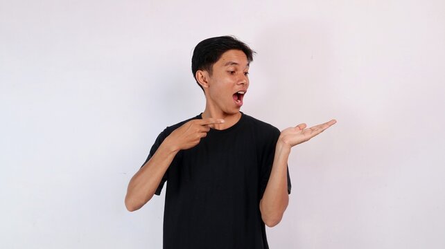 Young Asian man looking at his open palm in astonishment with an open mouth while pointing his finger toward it against a white background.