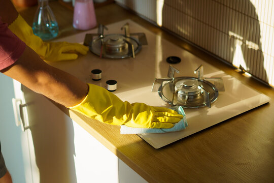 Close up of hands in yellow rubber gloves wiping a gas hob with a cloth, warm sunlight streaming in, ideal for household cleaning and hygiene concepts