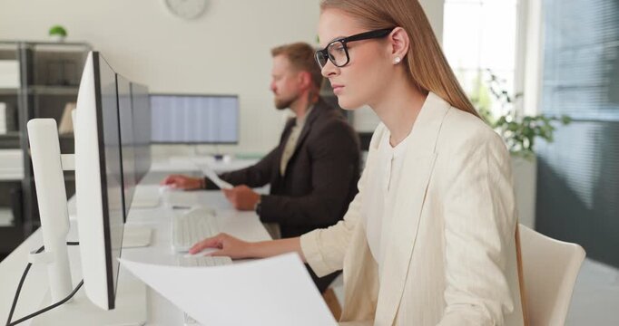 Accountant woman at computer in office reviewing report. She compares a document with database data on dual monitors, confirming entries as a colleague assists. Precise data analysis and focus.