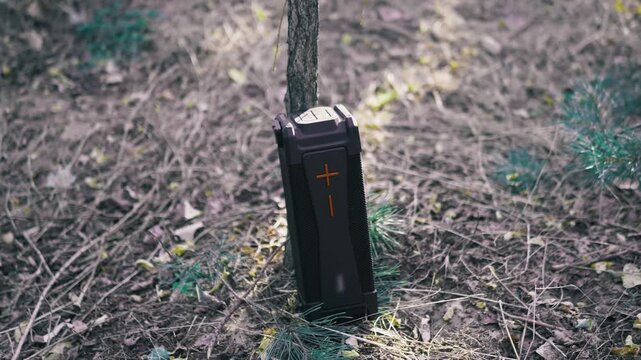 A black rugged portable speaker rests against a thin tree trunk on a wooded forest floor. This durable audio device delivers wireless sound for campers and hikers enjoying nature.