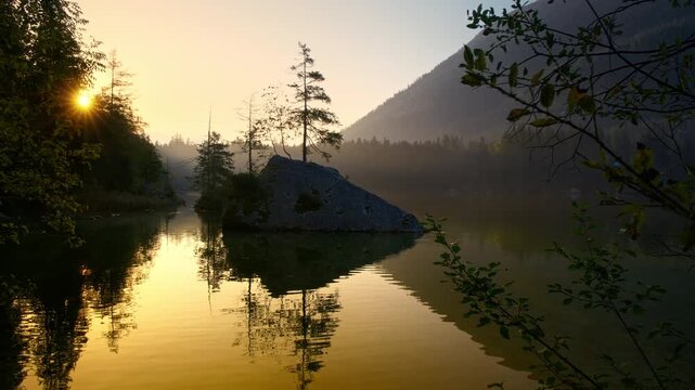 Golden sunrise at an idyllic lake in the German Alps. Tranquil slow panning shot with the silhouette of a small island on the gold water.