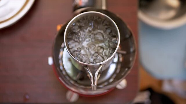 Water boils vigorously inside a stainless steel mug placed on a portable camping burner stove. An overhead perspective captures steam and bubbles during outdoor meal preparation.