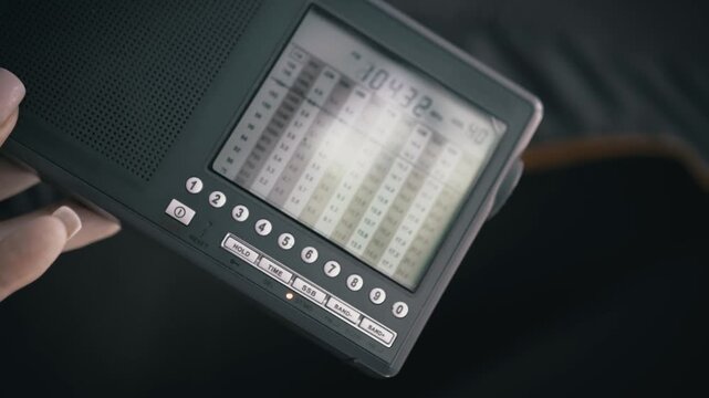 Hand holds portable shortwave radio showing illuminated digital frequency display screen. The electronic receiver features numbered control buttons and analog tuning scales.