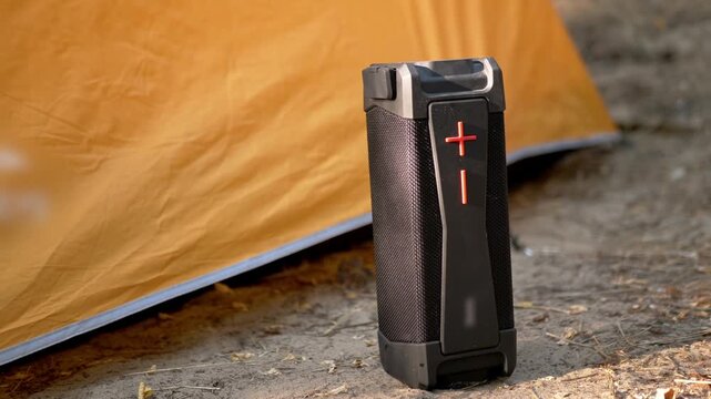 Black rugged portable speaker stands upright on the dry dirt beside an orange camping tent. The wireless device provides audio entertainment for outdoor adventures and leisure travel.