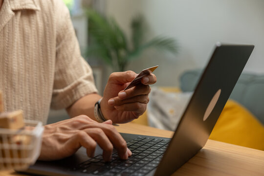 Adult hand using laptop keyboard and holding payment card during online shopping at home with warm interior light and focused financial activity