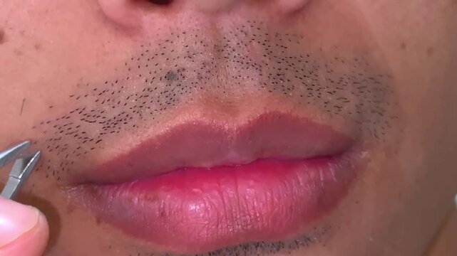 A young Asian man plucks his mustache and facial hair with metal tweezers at home for personal grooming and facial hygiene care.