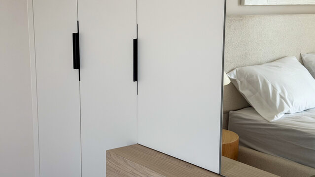 Modern neutral bedroom interior with built-in white wardrobe, large mirror, wooden furniture and cozy bed, contemporary minimalist home design.