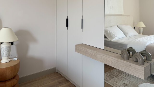 Modern neutral bedroom interior with built-in white wardrobe, large mirror, wooden furniture and cozy bed, contemporary minimalist home design.