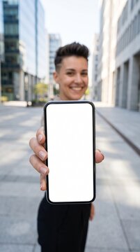 Vertical photo of a smiling woman holding a smartphone with a blank white screen. Wide angle perspective mockup of mobile phone in city street. Digital app presentation template