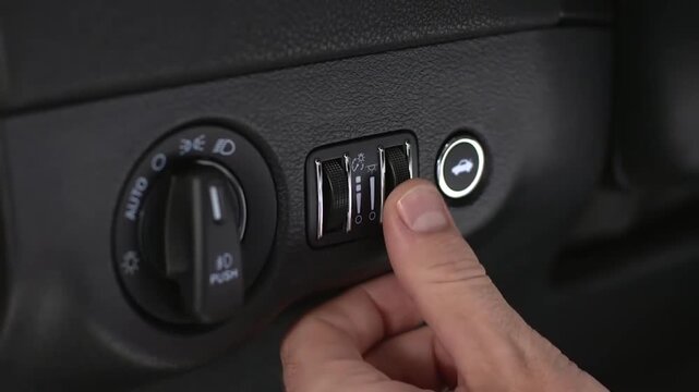 Close-up of hand pressing rocker switch on textured black dashboard panel inside modern car cockpit with ambient light during control operation and tactile grip detail