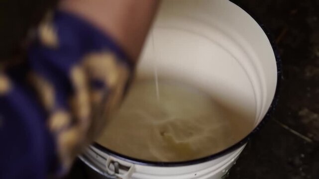 Hand in purple sleeve pours thick brown viscous liquid into white container at night, warm glow highlights textured ground and rim, soft-focus outdoor background