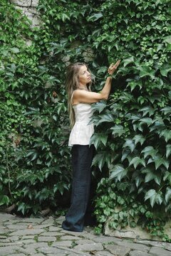 Middle-aged woman stands beside wall covered in green ivy in urban garden. She touches leaves with her hands, smiling at greenery. Sunlight shines through the foliage in background