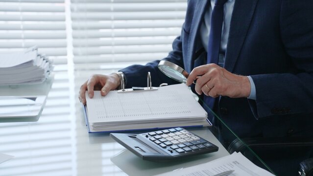 Professional auditor analyzing financial data with a magnifying glass, performing a detailed business investigation, checking documents, and verifying accounting records at the desk